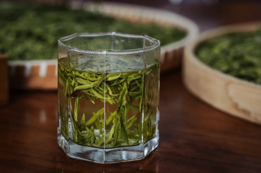 A glass of fresh green tea with vibrant leaves steeping in water sits on a wooden table. In the background are bamboo trays filled with more leaves.