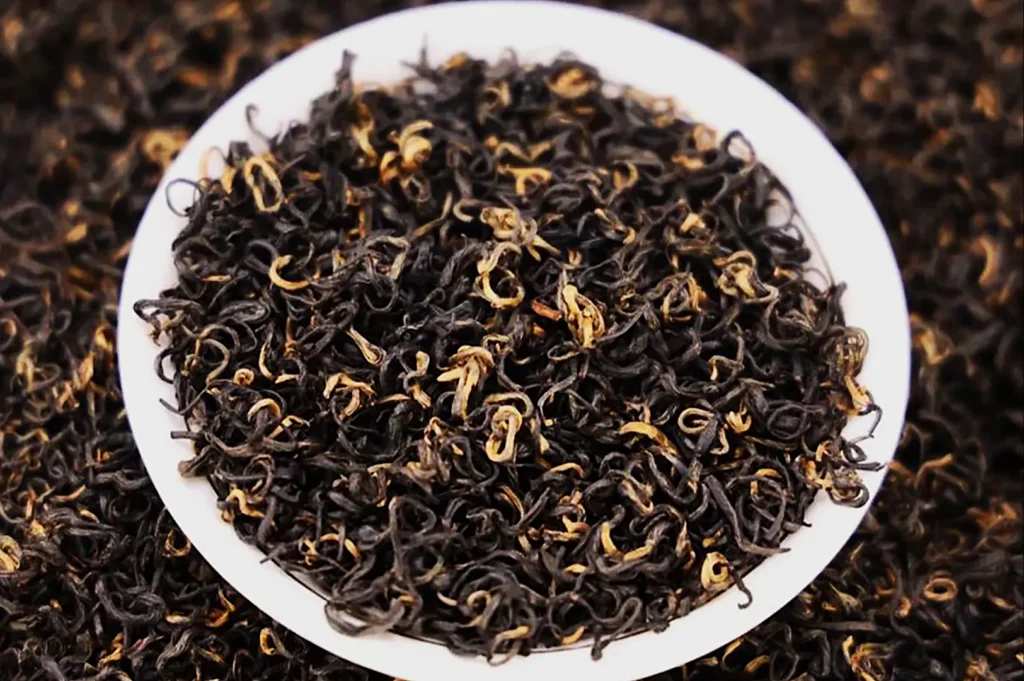 A close-up of a white plate filled with dried black tea leaves featuring a mix of dark and golden colors. The background shows more loose tea leaves.