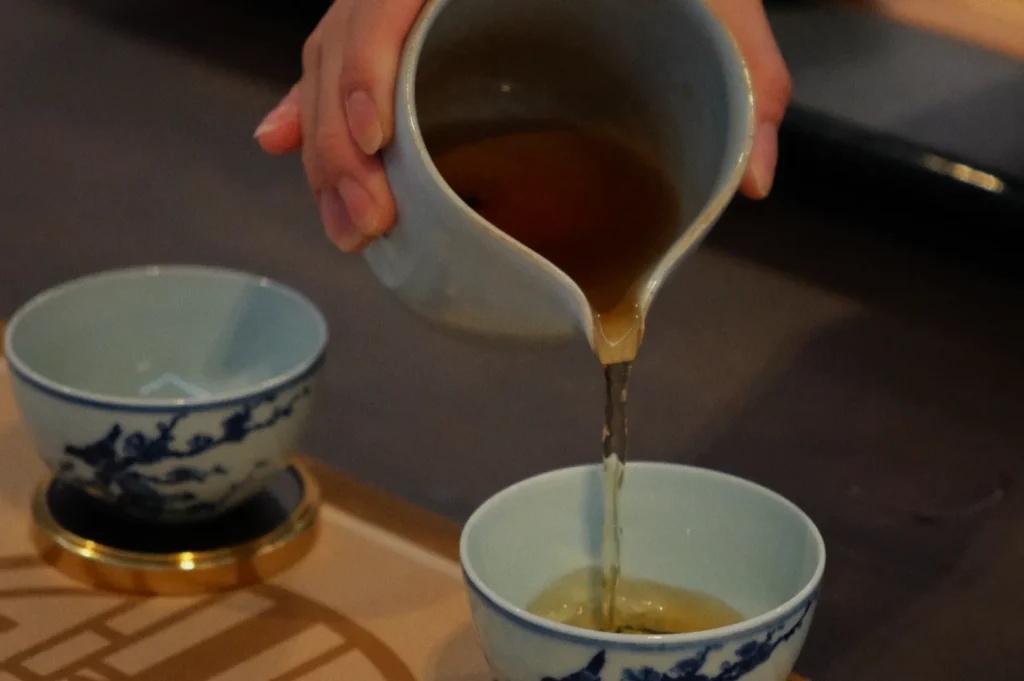 A hand pours tea from a spout into a delicate porcelain cup, adorned with a blue design. Another similar cup sits nearby, evoking a calm, ritualistic tone.