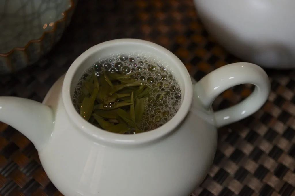 Ceramic teapot with green tea leaves floating in brew, bubbles on surface. Set on textured mat, evokes warmth and calm.
