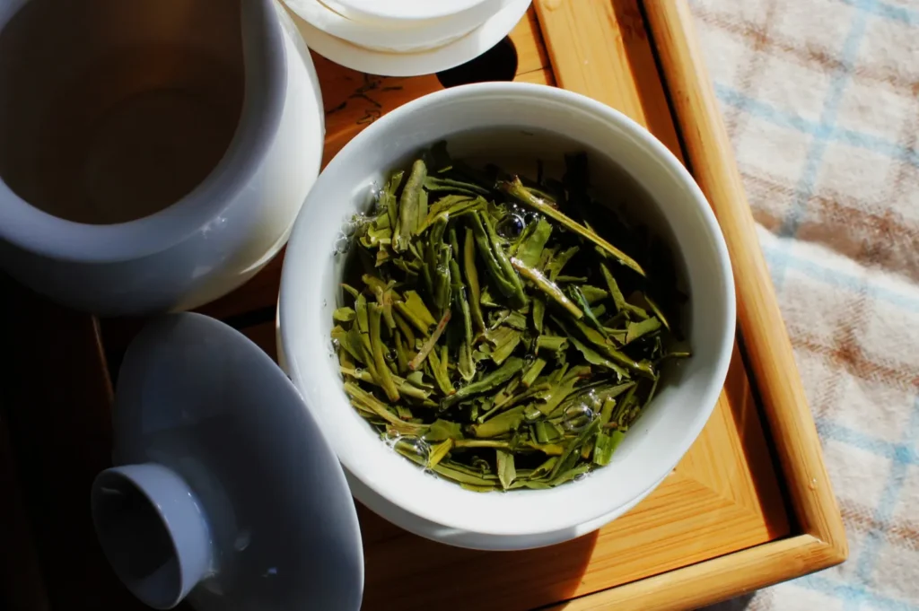 A white ceramic bowl filled with steeped green tea leaves sits on a bamboo tray, surrounded by other teaware. Warm sunlight highlights the scene.