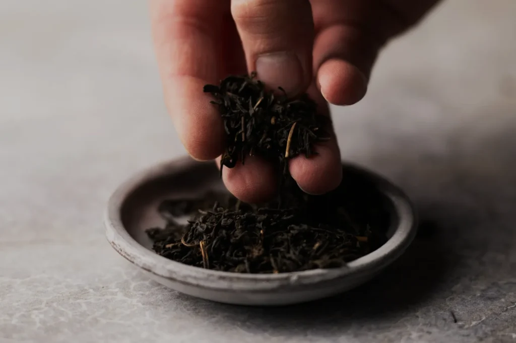 A hand holds loose black tea leaves over a small ceramic dish on a textured gray surface, conveying a sense of calm and simplicity in tea preparation.