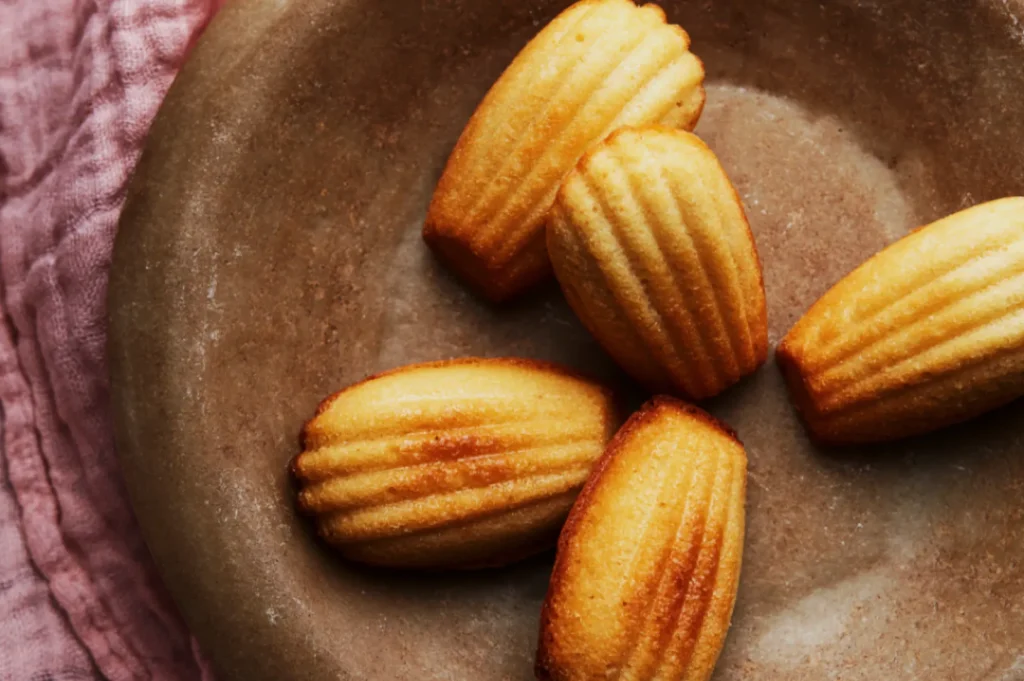 Golden madeleines with shell-like ridges are arranged on a brown plate, set atop a soft pink fabric. The scene feels warm and inviting.
