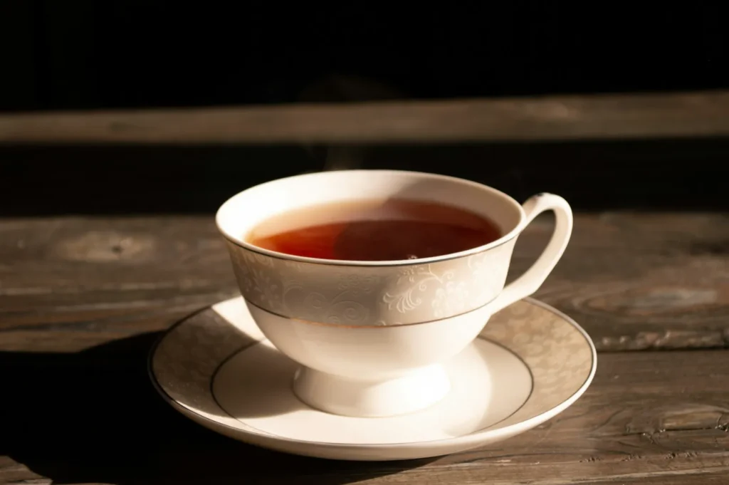 A steaming cup of tea sits in an elegant, white, floral-patterned teacup and saucer on a wooden table, bathed in warm sunlight.