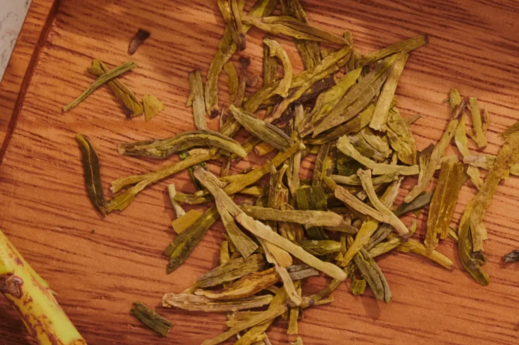 Dried green tea leaves scattered on a wooden surface, revealing earthy green tones and textures. The setting evokes a sense of calm and tradition.