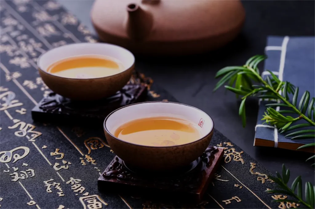 Two ceramic teacups filled with amber tea sit on ornate coasters atop a black mat with gold calligraphy. A teapot and greenery create a serene ambiance.