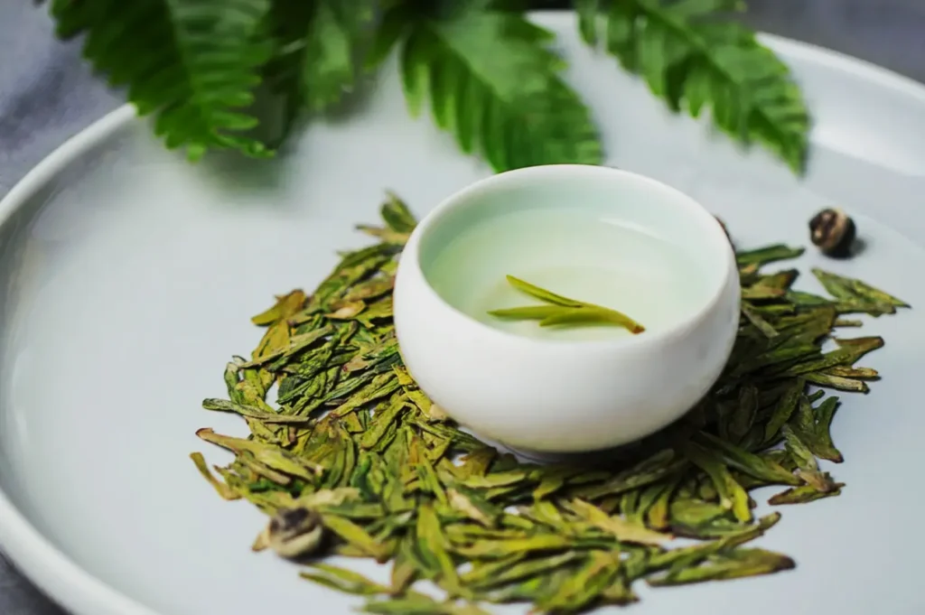 A white cup of light green tea sits on a plate surrounded by dried tea leaves, with a sprig of fresh leaves in the background. Calm and refreshing.