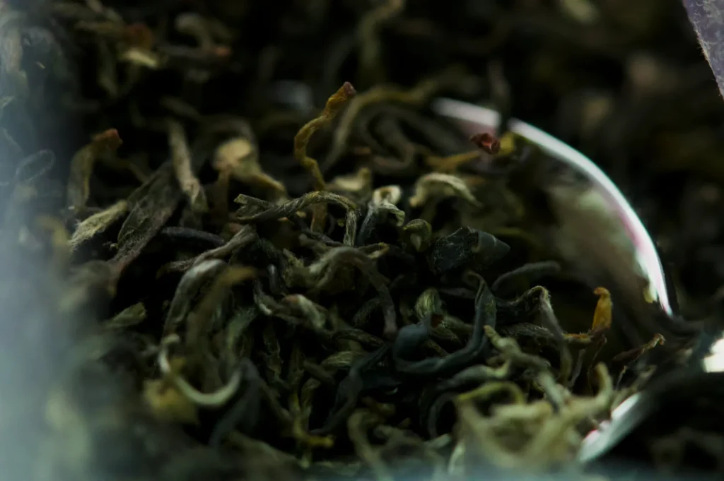 Close-up of loose dried tea leaves with curled edges, presenting a mix of dark green and brown hues. A silver spoon peeks from the right, adding a touch of shine.