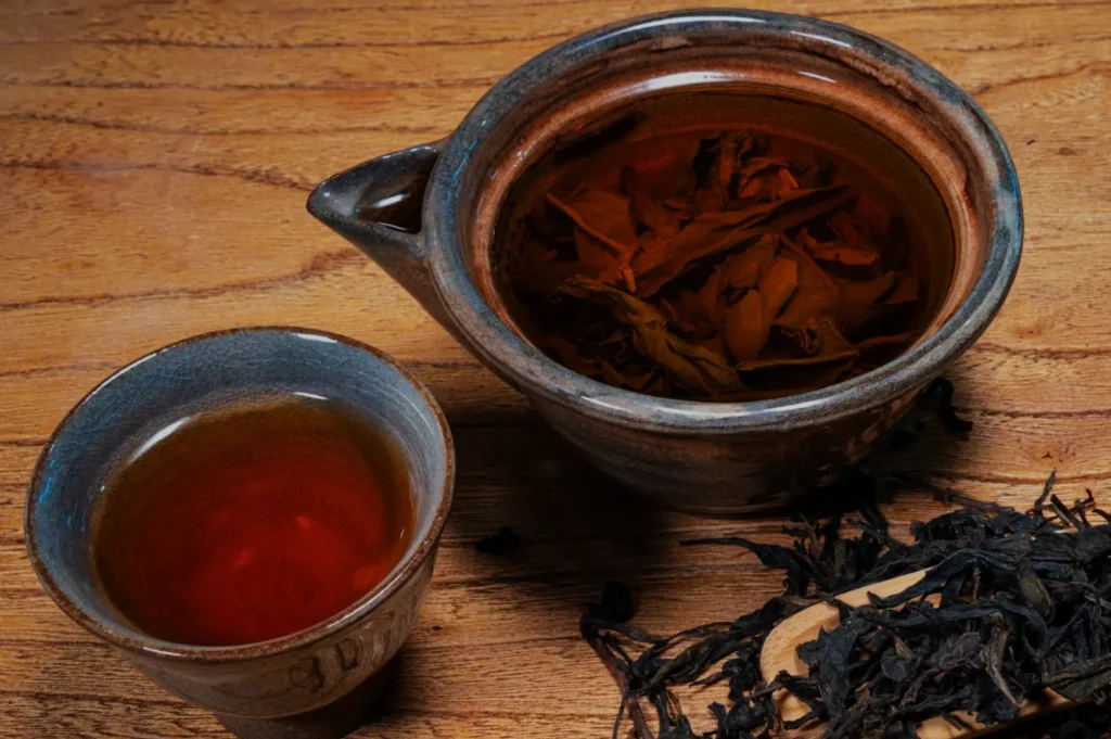 A rustic ceramic teapot filled with brewed tea leaves and a matching cup with amber tea sit on a wooden table. Loose tea leaves scattered nearby.