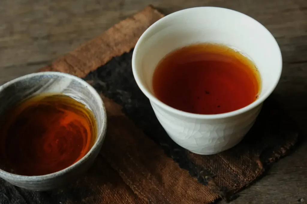 Two ceramic cups filled with dark amber tea sit on a rustic wooden surface with a brown cloth. The scene conveys warmth and simplicity.