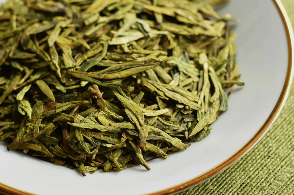 A close-up of dry green tea leaves on a white plate with a gold rim, set against a textured green cloth background, conveying freshness.