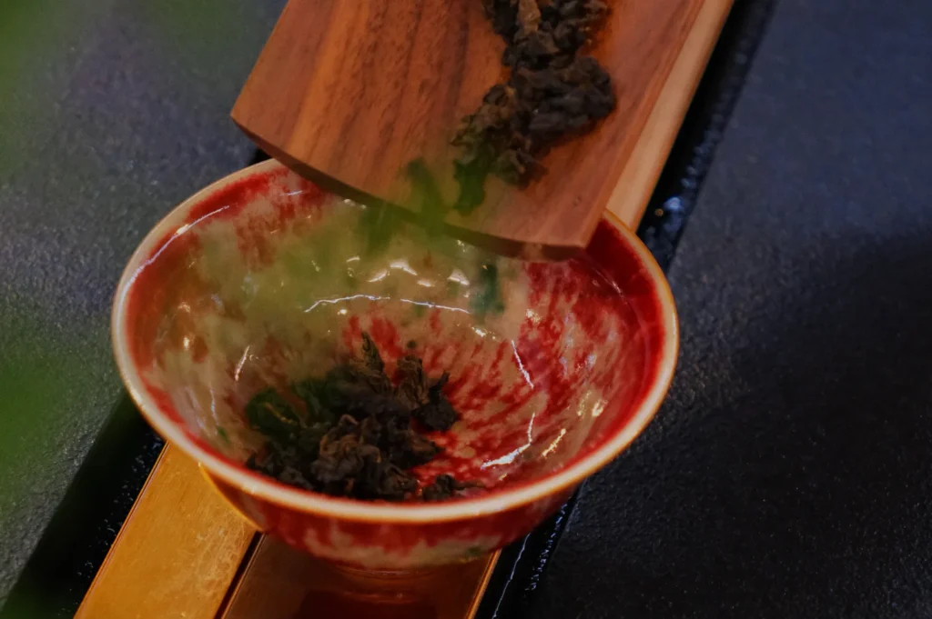 Loose tea leaves are being poured from a wooden scoop into a red and white ceramic bowl, creating a calming and traditional tea preparation scene.