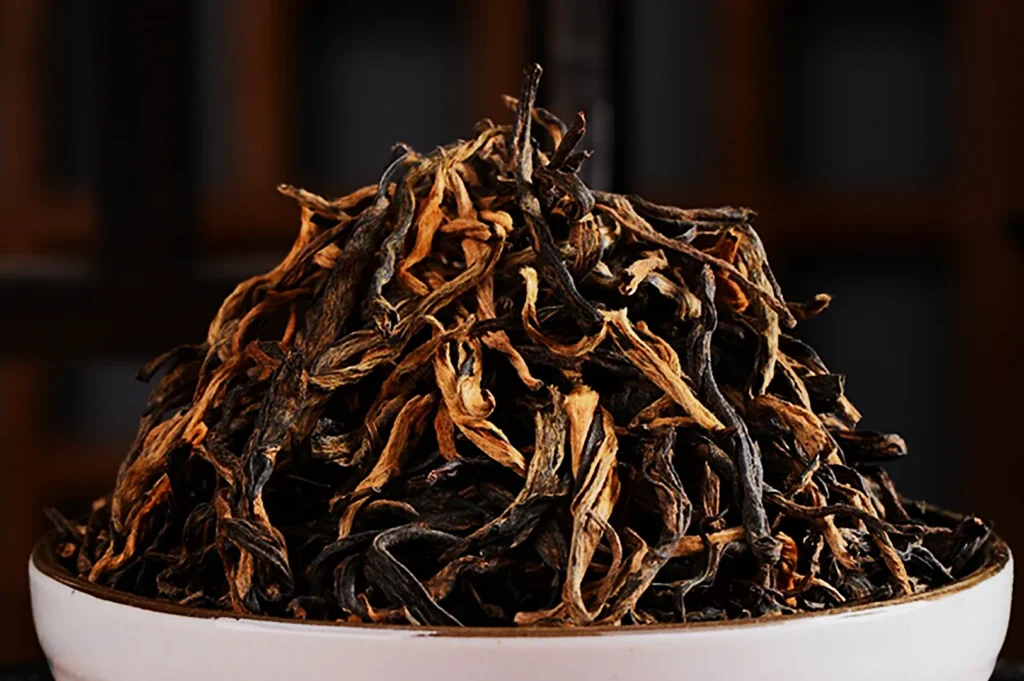A close-up of a white bowl filled with dry, twisted tea leaves in shades of brown and black, set against a blurred, dark wooden background.
