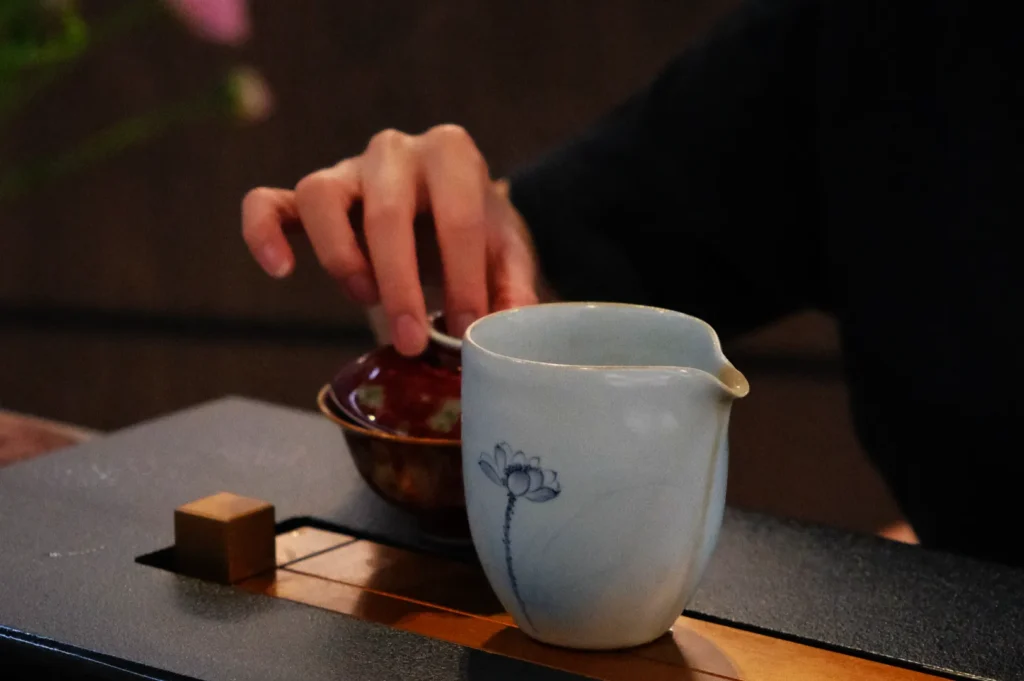 A hand reaches for a small red lidded bowl beside a white ceramic pitcher with a lotus design, set on a dark wooden tray. The scene indicates a peaceful, contemplative atmosphere.