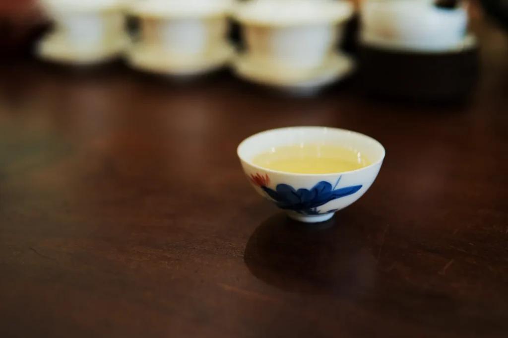 A small porcelain cup with floral designs, filled with light tea, sits on a smooth wooden table. Blurred white teacups and saucers are in the background.