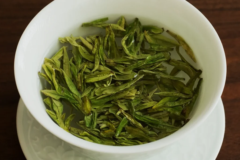 A white cup filled with hot water and loose, green tea leaves, creating a calming and natural tone. The cup rests on a wooden table.