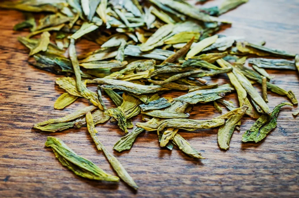 Loose green tea leaves spread across a wooden table, showcasing a mix of vibrant green and brown shades. The tone is natural and inviting.