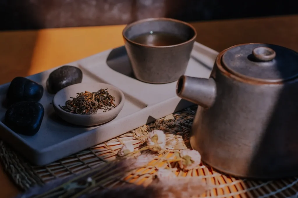 Ceramic teapot and cup with tea on a sunlit tray, alongside dark stones and dried flowers on a woven mat, creating a calm, warm ambiance.