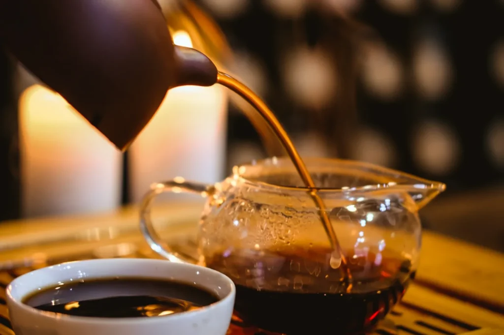 Tea is being poured from a brown pot into a glass teapot beside a cup of tea. The warm candlelight creates a cozy and inviting atmosphere.