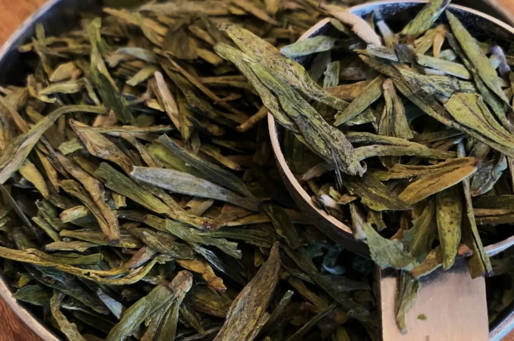 A plate of fresh green tea leaves on a bamboo mat, with a cardboard box labeled "Longjing Green Tea" in the background, conveying a sense of freshness.