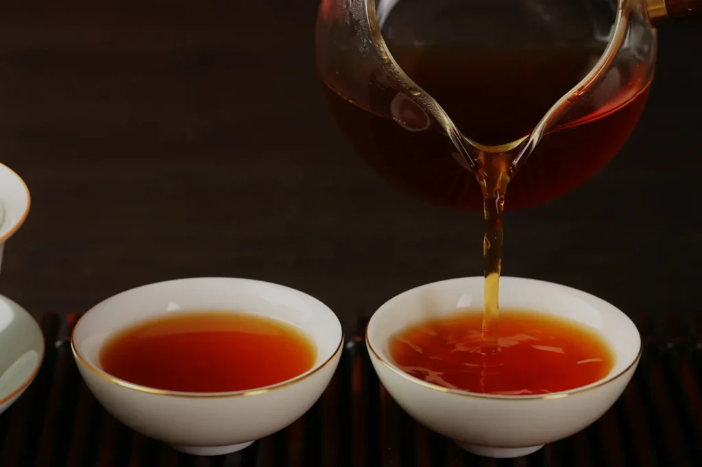 Tea is poured from a glass teapot into a white porcelain cup, filling it with rich amber liquid. The scene is calm and inviting.