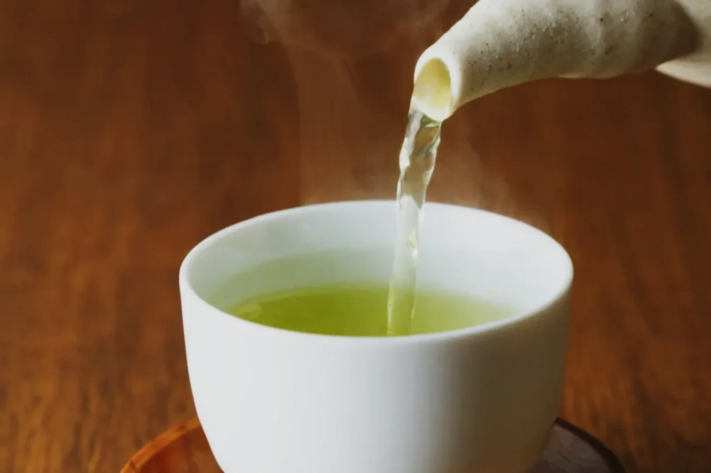 A steaming stream of green tea is being poured from a beige teapot into a white cup on a wooden surface, creating a warm and calming atmosphere.
