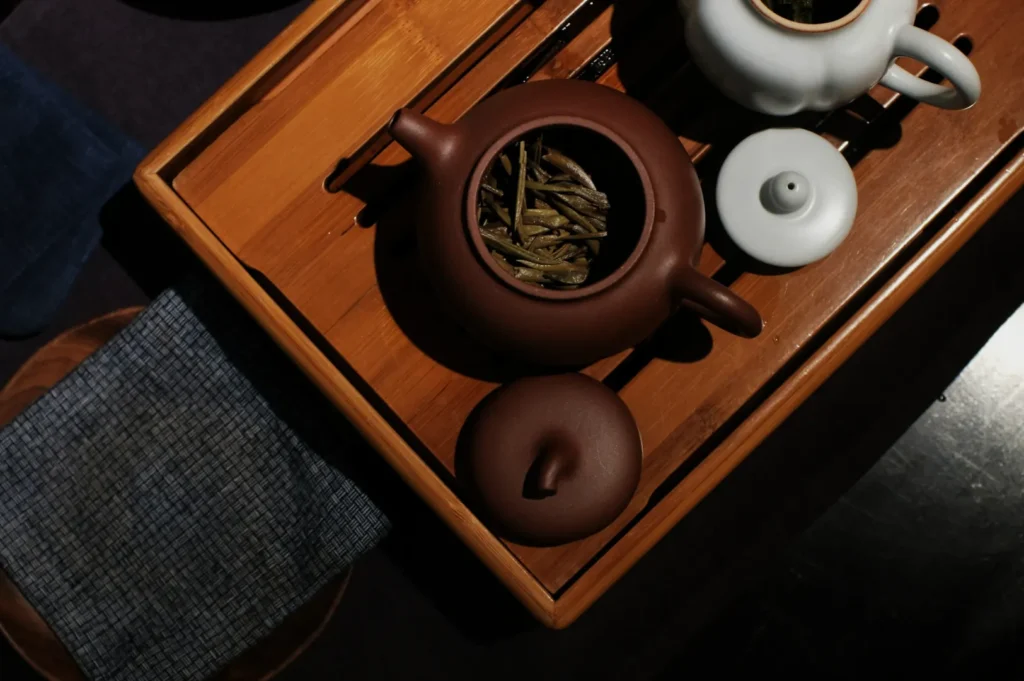 A brown clay teapot with tea leaves and a white ceramic teapot sit on a bamboo tray, creating a serene and traditional tea setting.