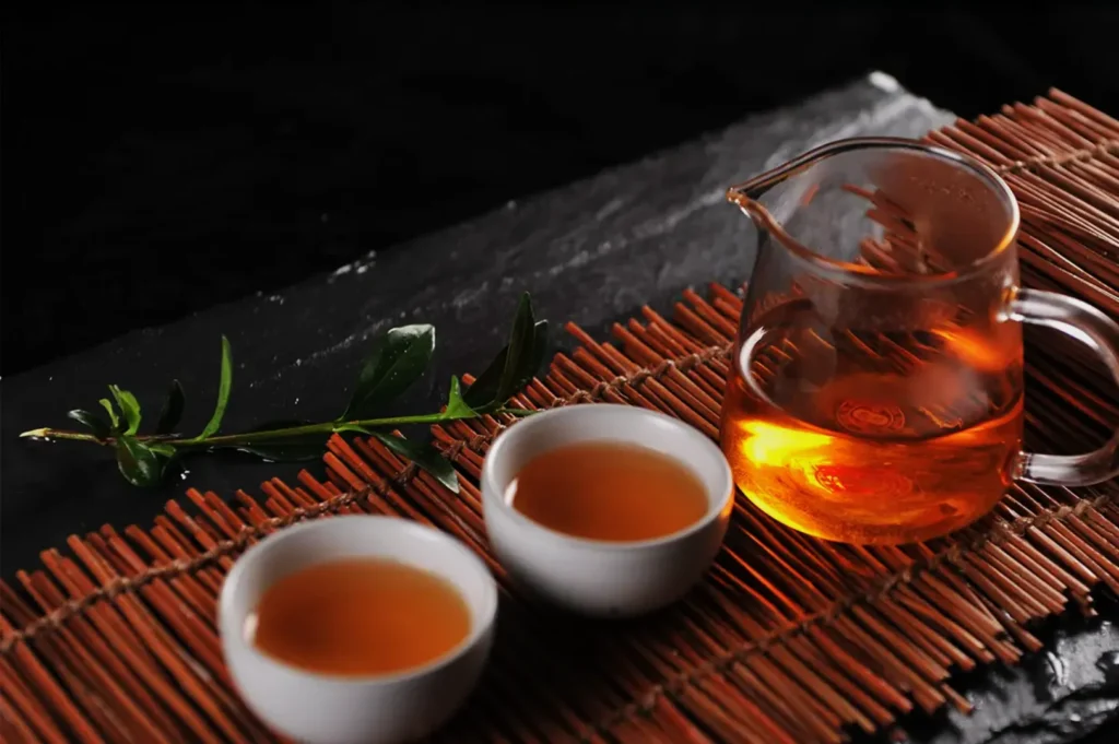 Warm tea in a glass pitcher and two cups on a brown bamboo mat, accompanied by a green leafy twig on a slate surface, creating a calming ambiance.