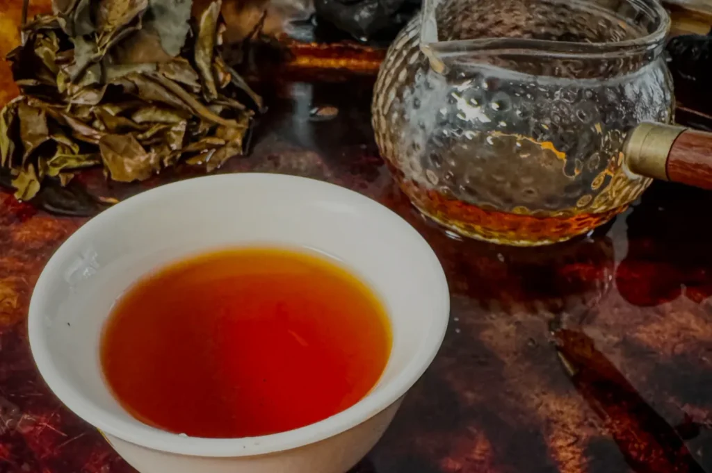 A steaming cup of reddish-brown tea in a white ceramic cup beside a textured glass teapot and dried tea leaves on a dark wooden surface.