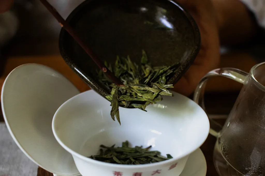 Tea leaves are being poured into a white gaiwan from a dark bowl using chopsticks. A glass teapot is partially visible in the background.