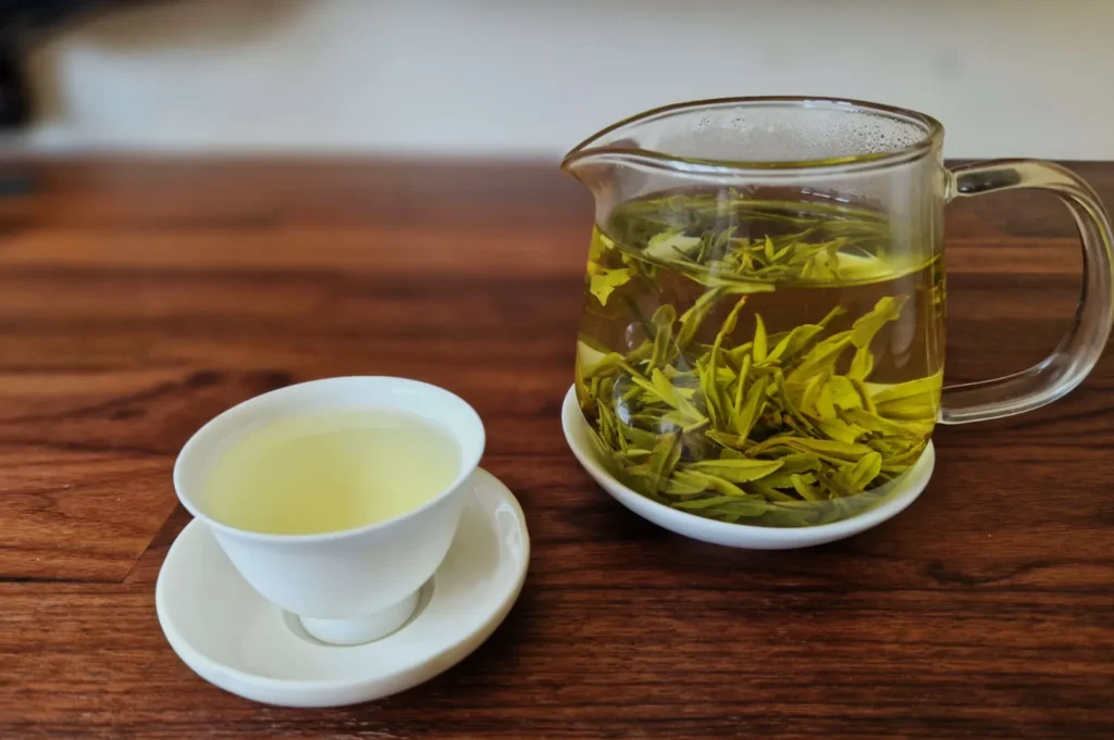 A clear glass teapot filled with green tea leaves and a white cup with light green tea sit on a wooden table, conveying calm and relaxation.