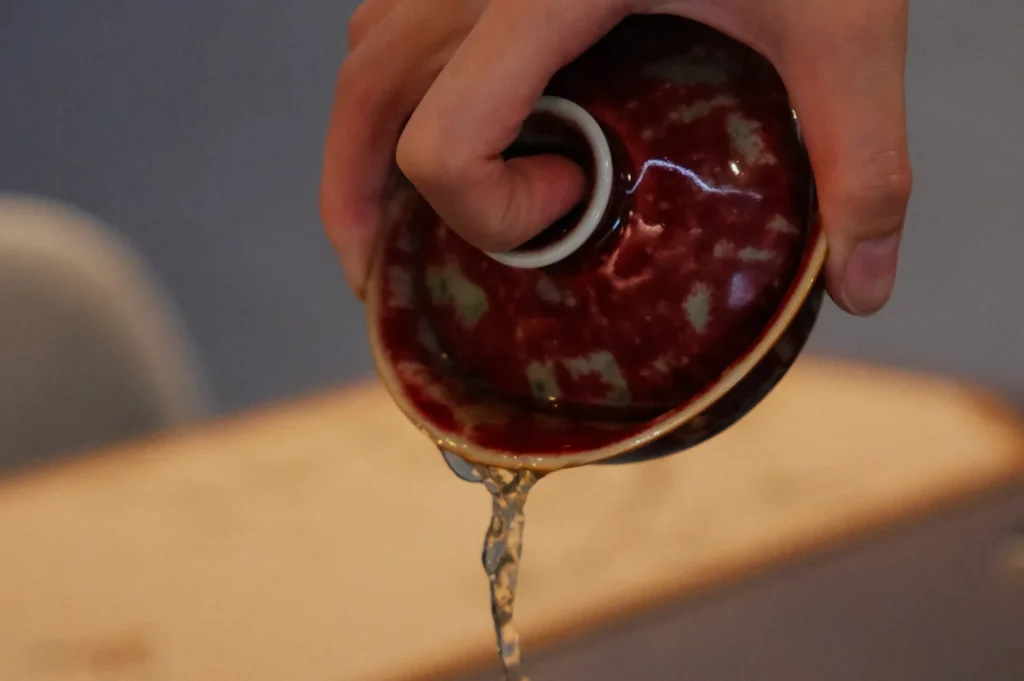 A hand carefully pours clear liquid from a small, red, textured ceramic bowl. The background is softly blurred, emphasizing the serene action.