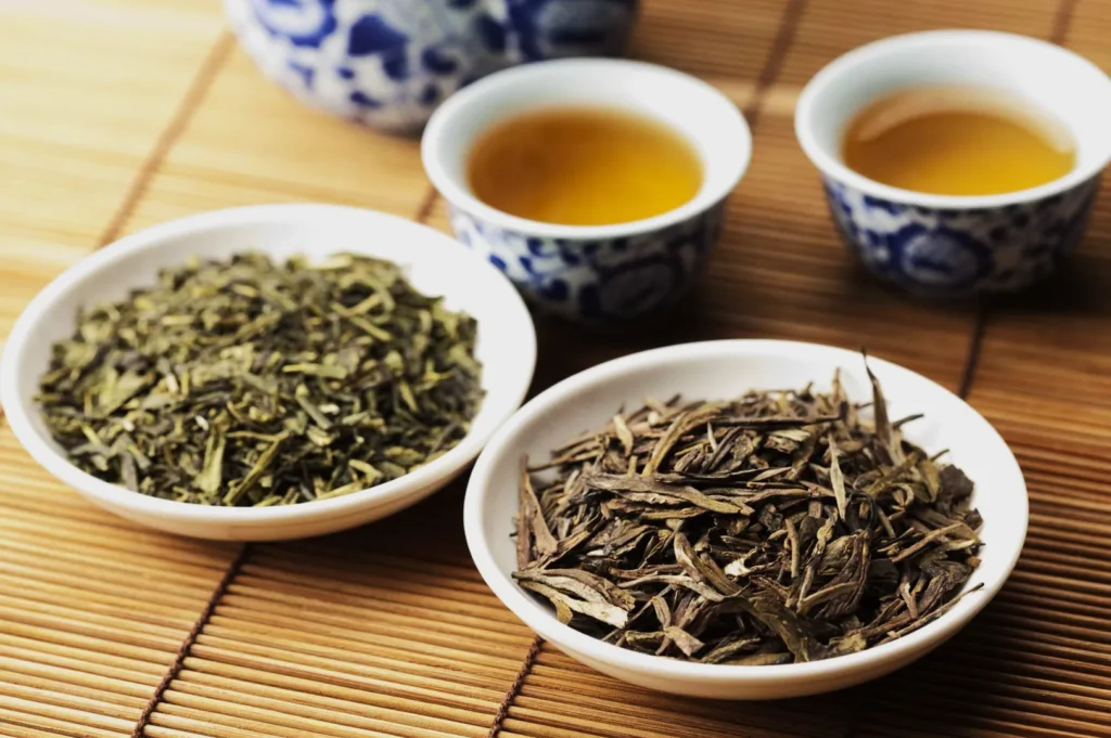 Two white bowls on a bamboo mat hold different loose tea leaves. In the background, two ornate cups contain brewed tea, creating a warm, inviting scene.