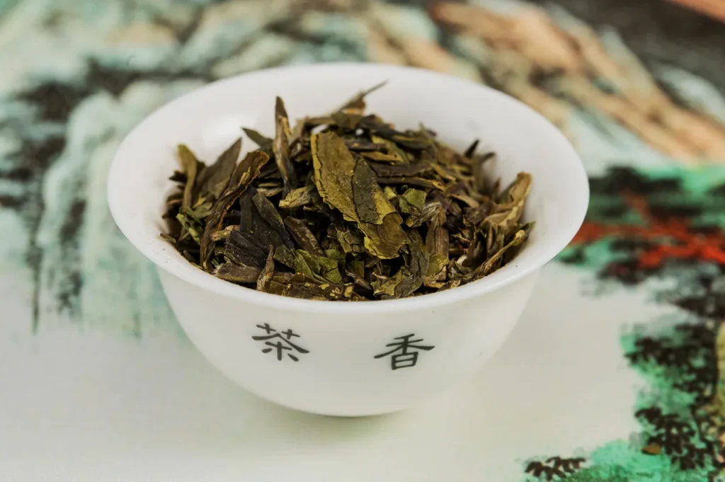 A small white bowl filled with loose green tea leaves sits on a colorful, blurred background. The bowl has Chinese characters printed on it, conveying an elegant, traditional atmosphere.