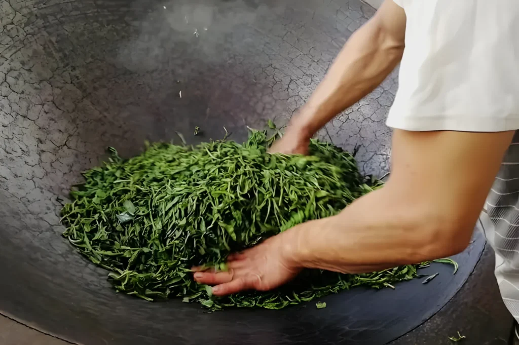 Person in a white shirt hand-roasting fresh green tea leaves in a large, cracked, black wok, conveying a sense of traditional craftsmanship.