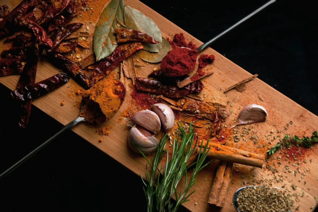A wooden board with various spices and herbs, including red chili peppers, garlic cloves, rosemary, bay leaves, and cinnamon sticks, against a dark background.
