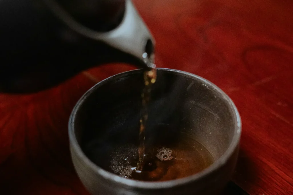 Pouring steaming tea into a dark cup on a rich wooden table. The scene evokes warmth and relaxation through its cozy and inviting atmosphere.