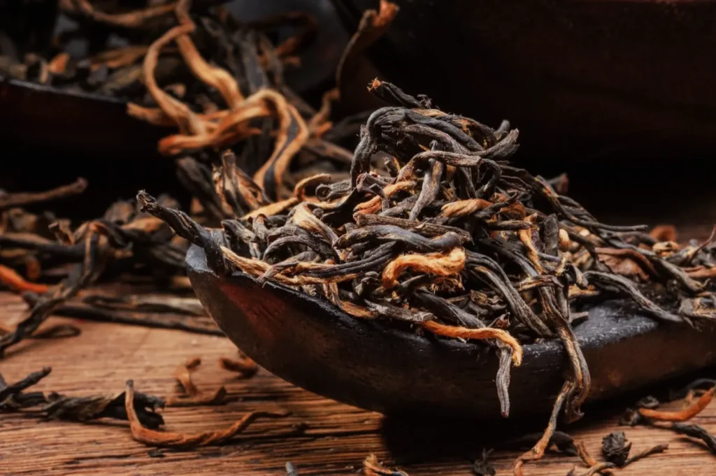 A close-up of loose black tea leaves with orange tips on a wooden surface. The dark, twisted leaves create a rustic and earthy atmosphere.