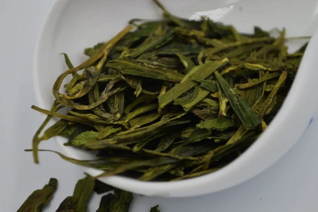 Close-up of dried green tea leaves in a white bowl on a light surface. The leaves are elongated and vibrant green, conveying freshness and aroma.