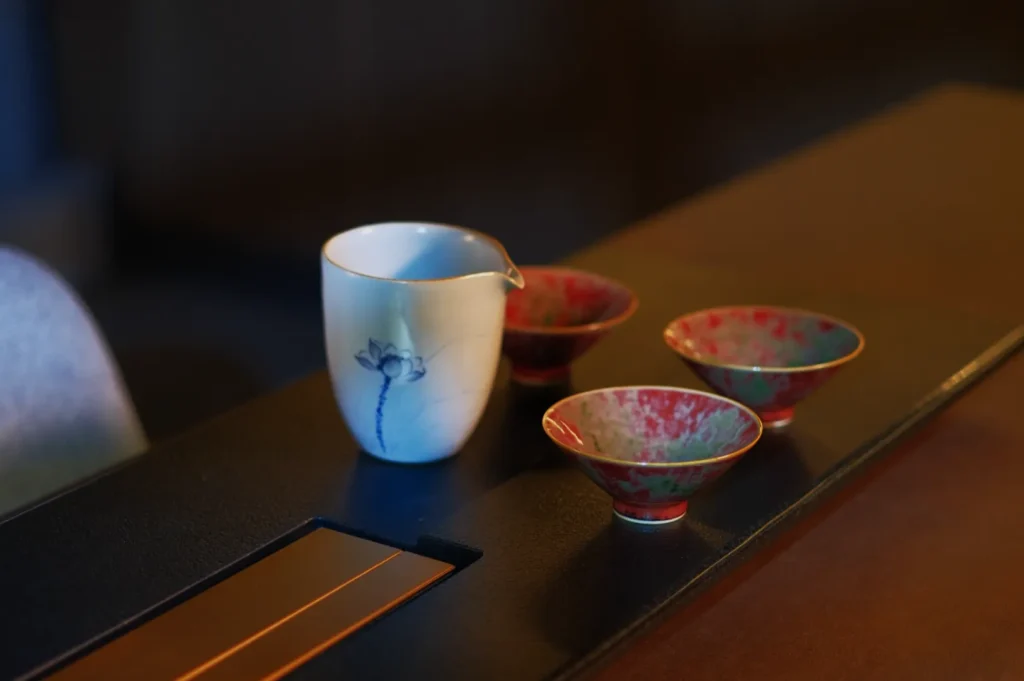 Ceramic pitcher with floral design and three speckled red-green cups on a wooden table. The setting is elegant and serene, evoking a warm ambiance.