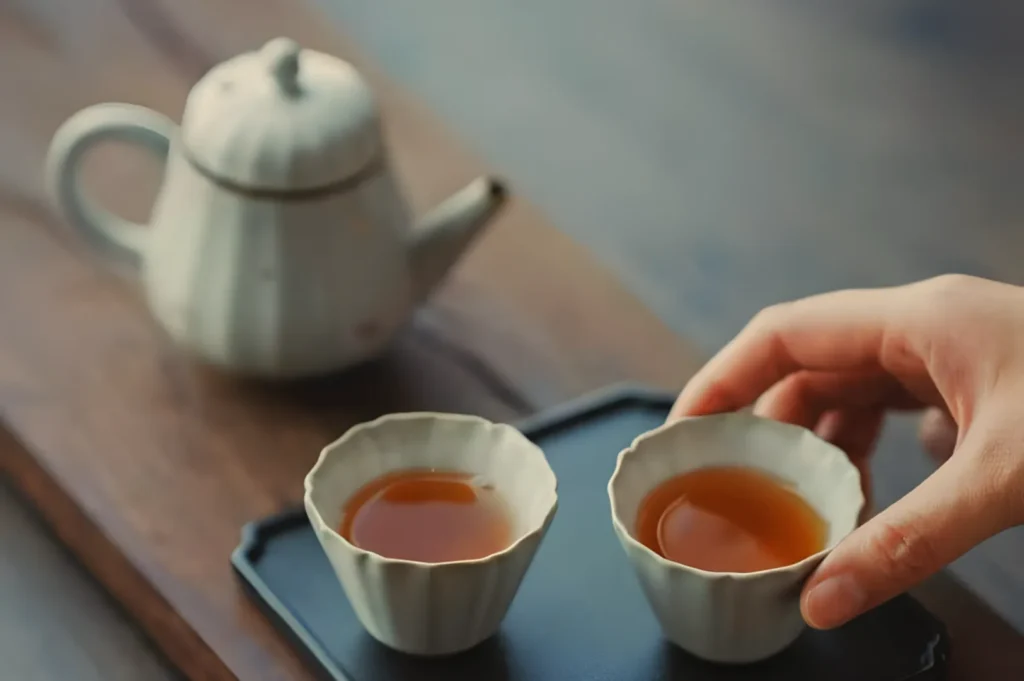 A hand reaches for one of two white ceramic tea cups filled with amber tea on a tray, with a matching teapot in the background on a wooden table.