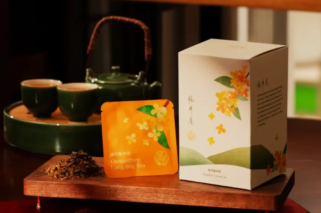 Packaging of Osmanthus Long-ling Tea on a wooden tray. Includes a tea box with floral design, a sachet, loose tea leaves, and green teapot set. Cozy ambiance.