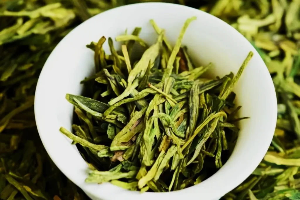 A white bowl filled with loose long leaves of green tea, placed on a background of more tea leaves. The image conveys freshness and vitality.