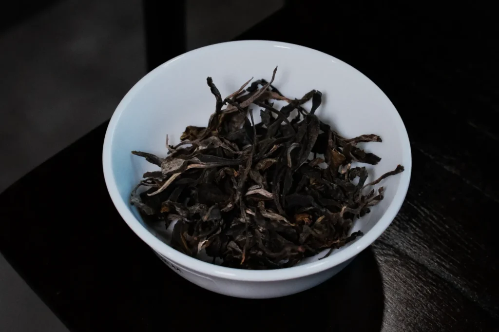 A white bowl filled with dry, twisted dark tea leaves sits on a dark wooden table. The setting exudes a calm, rustic tone, highlighting the tea's earthy quality.