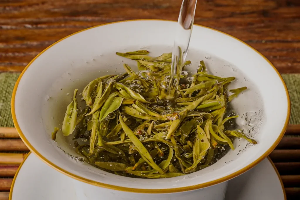 Pouring water into a white cup filled with green tea leaves, creating bubbles. The scene is warm and inviting, set on a wooden table with a calm ambiance.