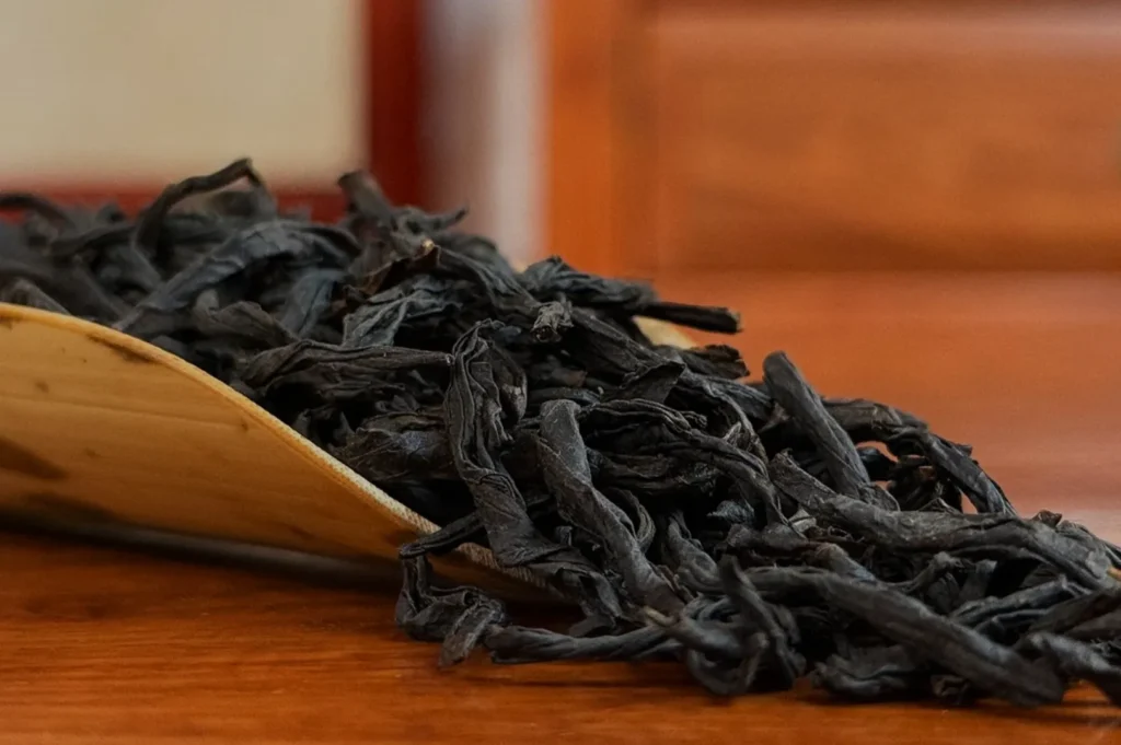 A pile of dark, twisted oolong tea leaves rests on a curved bamboo tray on a wooden table, evoking a rustic and organic feel against a blurred backdrop.