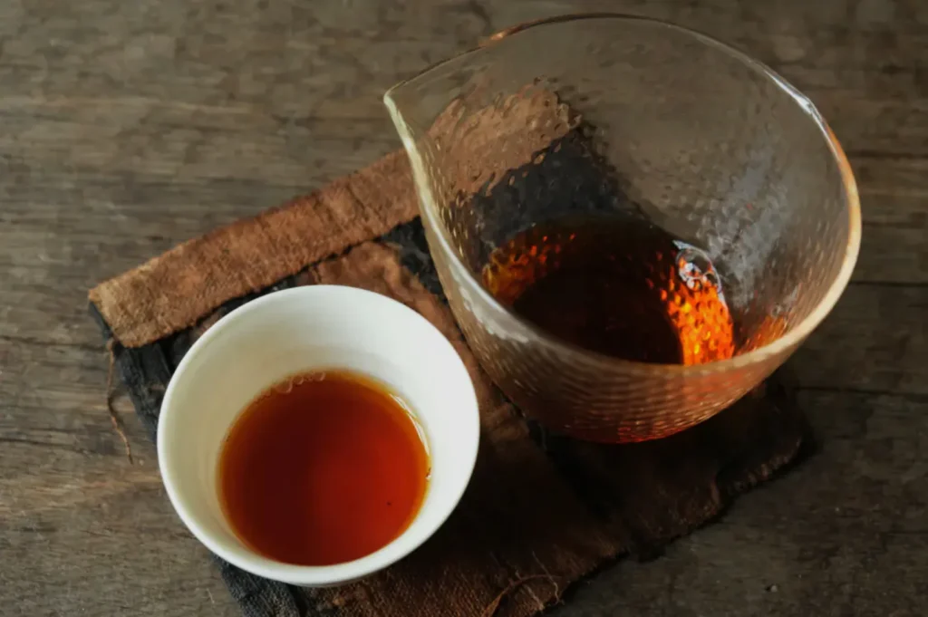A glass pitcher and white cup filled with amber tea sit on a rustic wooden surface with a brown cloth, conveying a warm, serene atmosphere.