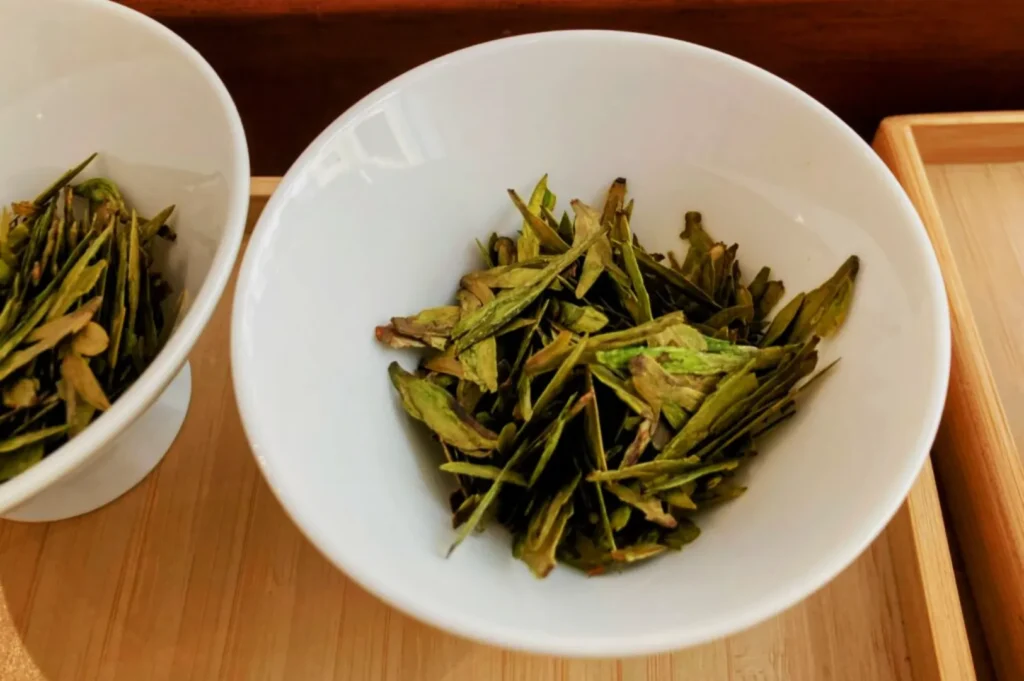 White bowls on a wooden tray, filled with vibrant green tea leaves. The scene conveys a fresh, natural, and serene atmosphere.