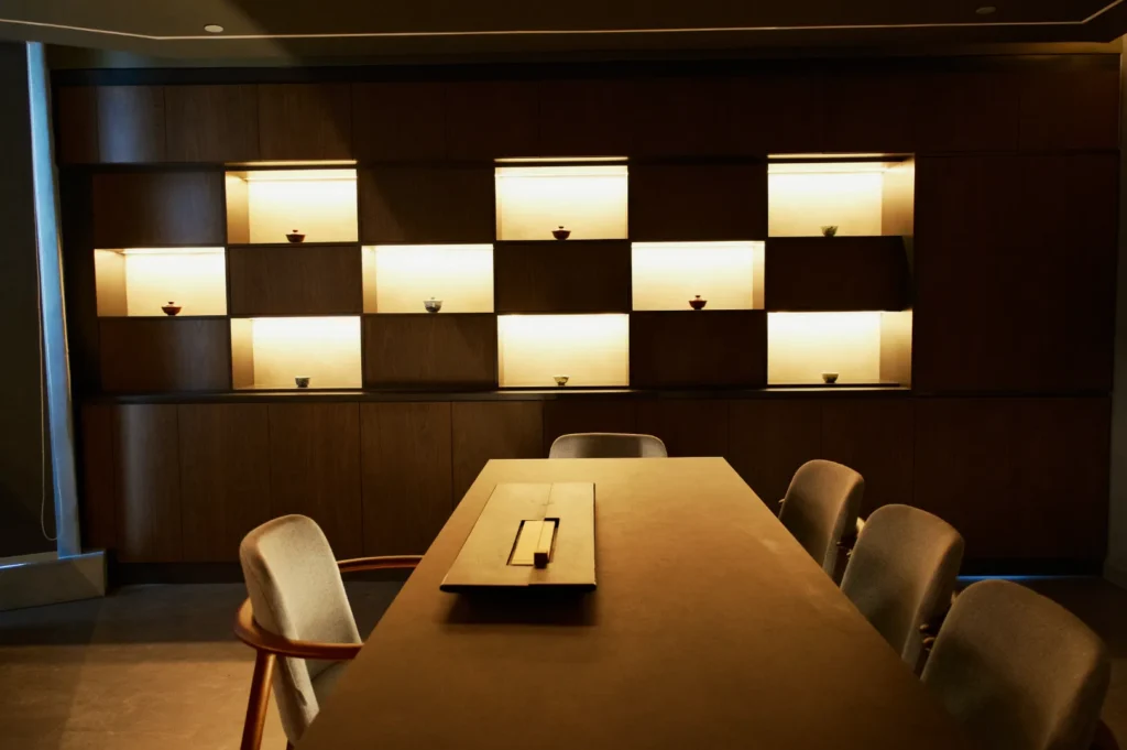 Modern conference room with a long wooden table surrounded by chairs. A backlit shelving unit displays small decorative bowls, creating a warm ambiance.