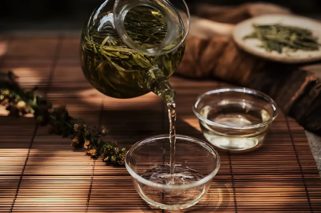 A glass teapot pours herbal tea into a small glass cup on a bamboo mat. Sunlight gently illuminates the scene, evoking a calm, serene atmosphere.